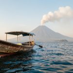 Bateau traditionnel indonésien naviguant sur une mer calme face au volcan Anak Krakatau, sous une douce lumière matinale et avec un léger panache au-dessus du cratère.