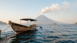 Bateau traditionnel indonésien naviguant sur une mer calme face au volcan Anak Krakatau, sous une douce lumière matinale et avec un léger panache au-dessus du cratère.
