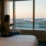 Une jeune voyageuse seule se repose dans une chambre d’hôtel près d’un aéroport à l’aube, avec sa valise et la piste éclairée visible par la fenêtre.
