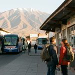 Un homme et une femme voyageurs portant des sacs à dos attendent devant un terminal de bus à La Paz, avec les montagnes de l’Altiplano en arrière-plan dans la lumière dorée du matin.