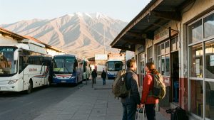 Un homme et une femme voyageurs portant des sacs à dos attendent devant un terminal de bus à La Paz, avec les montagnes de l’Altiplano en arrière-plan dans la lumière dorée du matin.